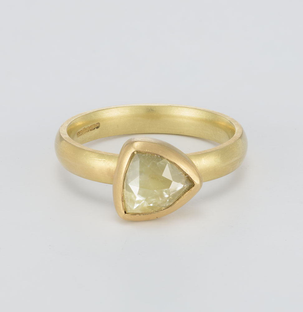 A yellow gold ring featuring a triangular-cut pale yellow gemstone in a bezel setting, shown on a light gray background.