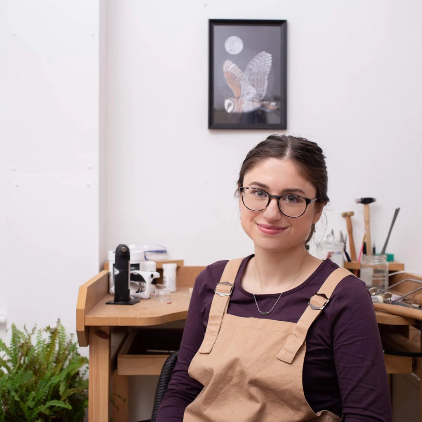 A woman with glasses and a brown apron over a dark purple shirt sits in a bright, modern art studio with wooden workbenches, tools, and framed wall art.